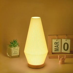 May include: A wooden table lamp with a white pleated shade. The lamp is turned on and illuminating the room. The lamp is sitting on a wooden surface next to a calendar that says "MAR 0".