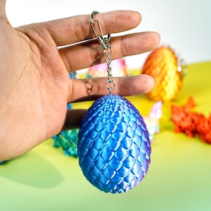 May include: A blue and purple dragon egg keychain with a silver chain and clasp. The egg has a textured, scale-like pattern. Other dragon egg keychains in orange, green, and red are visible in the background.