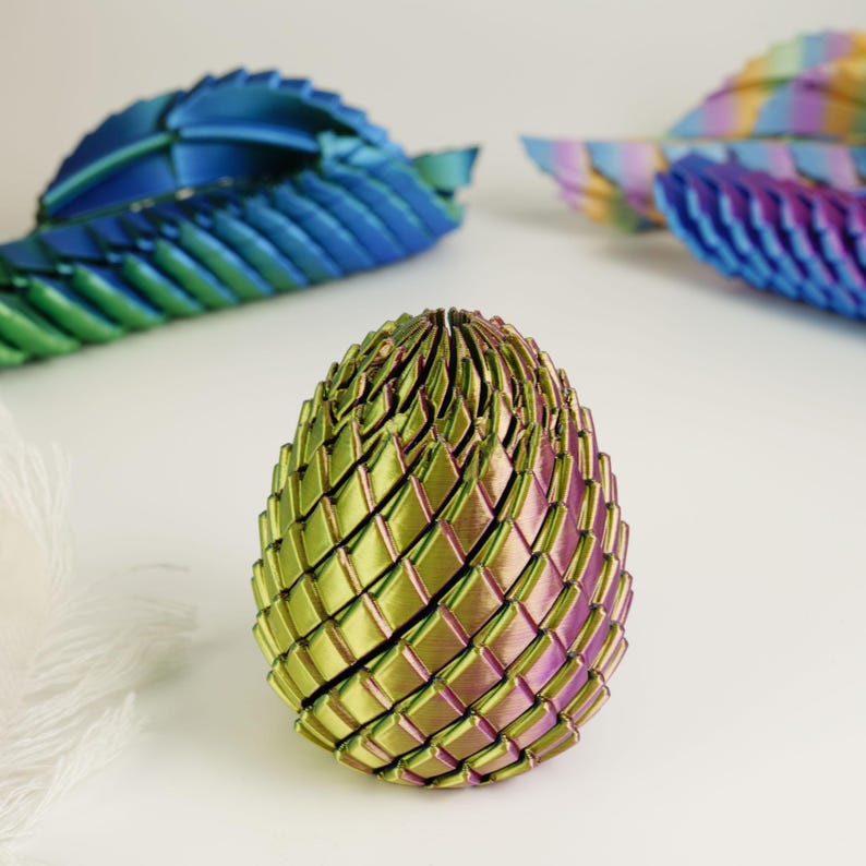 May include: A 3D-printed, iridescent egg-shaped sculpture with a woven pattern. The egg transitions from green to purple hues. Two similar, abstract leaf-like sculptures in blue and purple are in the background.