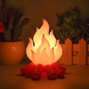 Pode incluir: Lâmpada decorativa em forma de fogueira com uma luz quente e brilhante. A lâmpada apresenta um design de chama em tons de laranja e amarelo, apoiada em troncos vermelho-alaranjados. A lâmpada foi projetada para se assemelhar a uma fogueira.