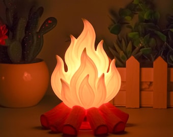 May include: A decorative campfire-shaped lamp with a warm, glowing light. The lamp features a flame design in shades of orange and yellow, resting on red-orange logs. The lamp is designed to resemble a campfire.