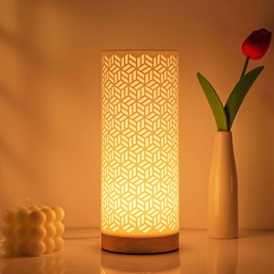 Puede incluir: Una lámpara de mesa cilíndrica con un patrón geométrico, que emite un brillo cálido. La lámpara se asienta sobre una base de madera. También hay un jarrón blanco con un tulipán rojo y una vela de burbujas blanca.