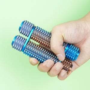 May include: Two blue and brown massage rollers with a textured, triangular pattern are held in a hand against a light green background. The rollers have blue ends and a ridged design.