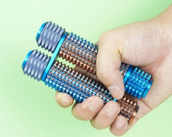 Könnte beinhalten: Zwei blaue und braune Massagerollen mit einem strukturierten, dreieckigen Muster werden in einer Hand vor einem hellgrünen Hintergrund gehalten. Die Rollen haben blaue Enden und ein geriffeltes Design.