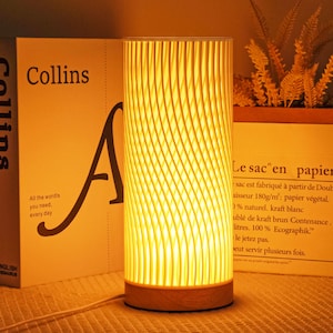 Pode incluir: Uma luminária de mesa cilíndrica com um brilho amarelo quente. A luminária tem uma base de madeira e uma tela texturizada e canelada. A luminária está ao lado de um livro com a palavra "Collins" na capa.