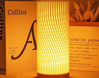 May include: A cylindrical table lamp with a warm, yellow glow. The lamp has a wooden base and a textured, ribbed shade. The lamp is next to a book with the word "Collins" on the cover. The text "All the wordis you need, every day" is also visible.