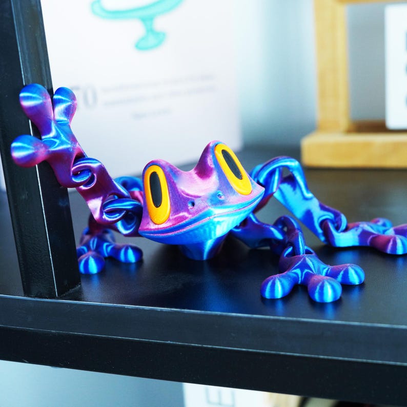 May include: A vibrant, multi-colored frog figurine with a glossy finish. The frog features a blend of blue, purple, and pink hues, with yellow eye accents. The frog is posed on a dark surface, showcasing its detailed design and playful aesthetic.