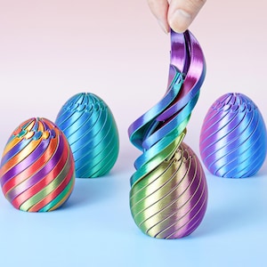 Può includere: Una collezione di sculture di uova di Pasqua iridescenti dai colori metallici. Un uovo viene manipolato per mostrare il suo design flessibile a forma di nastro. Le uova sono nelle tonalità di viola, blu, verde, oro e arancione, con un motivo a spirale.