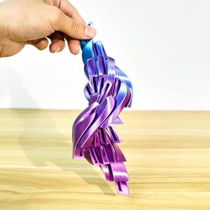 May include: A hand holds a 3D-printed abstract sculpture. The sculpture is in shades of purple and blue, with a layered, ribbon-like design and a metallic sheen. The object is displayed against a light background.