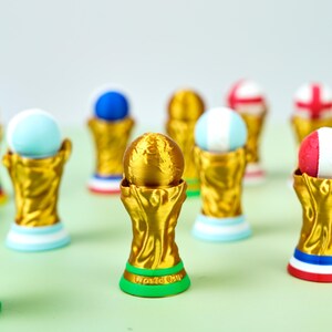Könnte beinhalten: Eine Sammlung von Miniatur-Trophäenfiguren in Gold, jede mit einem Globus oben und einem farbigen Sockel. Die Sockel tragen den Text "World Cup". Die Globen sind in verschiedenen Farben bemalt, darunter Blau, Rot und Weiß.