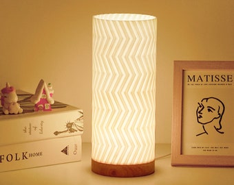 May include: A cylindrical table lamp with a white, zig-zag patterned shade and a wooden base. The lamp is illuminated, casting a warm glow. Decorative items and a framed print are visible in the background.