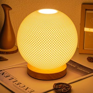 May include: A spherical table lamp with a woven, grid-like shade, emitting a warm, yellow glow. The lamp sits on a round, wooden base. A brown vase and a framed picture are in the background. The lamp is a decorative lighting fixture.