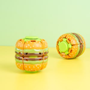 May include: Two plastic fidget toys shaped like hamburgers. Each toy features stacked layers of orange bun with white sesame seed details, green lettuce, and a brown patty. The toys have a segmented, mechanical design with a central green circular component.
