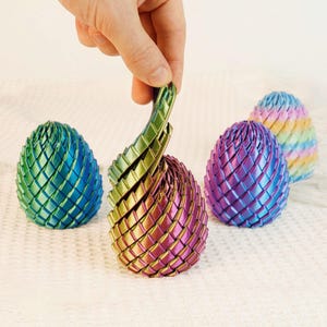 May include: A hand holds a section of a metallic, multi-colored egg-shaped sculpture. The sculpture is made of interwoven strips, with a gradient of colors including green, gold, and purple. Other similar sculptures in blue, purple, and rainbow hues are in the background.