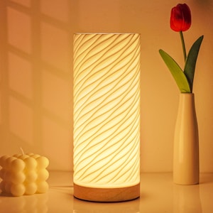 Puede incluir: Una lámpara de mesa cilíndrica con un patrón ondulado, que emite un brillo cálido. La lámpara tiene una base de madera y está junto a un jarrón blanco con un tulipán rojo. También hay una vela amarilla con forma de burbuja.