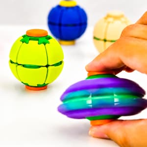 May include: A collection of spherical fidget toys featuring segmented panels. One toy is lime green with orange caps, another is blue with yellow caps, one is cream-coloured, and a purple and teal gradient toy is held in the foreground.