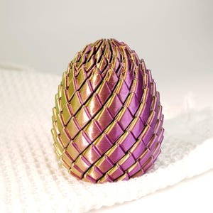 May include: A decorative egg-shaped object with a woven design. The egg features a gradient of colors, transitioning from green to purple. The object is resting on a white, textured surface. The egg's intricate design and color scheme create a visually appealing aesthetic.