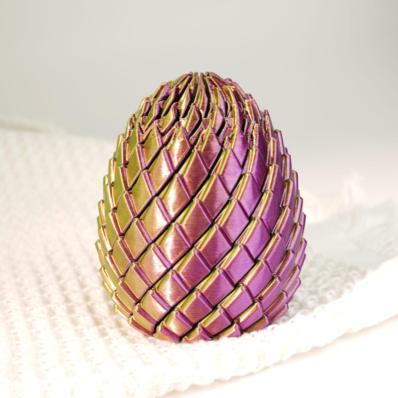 May include: A decorative egg-shaped object with a woven design. The egg features a gradient of colors, transitioning from green to purple. The object is resting on a white, textured surface. The egg's intricate design and color scheme create a visually appealing aesthetic.