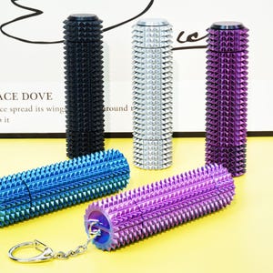May include: A collection of metal massage rollers and keychains in various colors, including black, silver, blue, and purple. Each roller features a studded design. The keychains are attached to the rollers with a small metal ring.