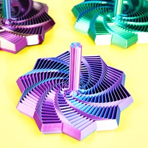 Puede incluir: Tres coloridas peonzas geométricas en tonos morado, azul y verde. Las peonzas tienen un diseño en espiral y están sobre un fondo amarillo brillante. La peonza central está enfocada, mostrando su intrincada construcción en capas.