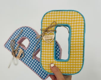 May include: Two fabric letters, an "O" and an "R", are displayed. The "O" is yellow and white gingham with a blue border. The "R" is blue and white gingham with a pink border. Both have wooden heart tags with names.
