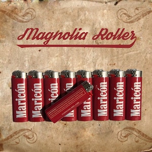 May include: A group of ten red lighters with the word "Maricón" printed on each one. The lighters are arranged in a row on a brown surface. The text "Magnolia Roller" is printed in red at the top of the image.