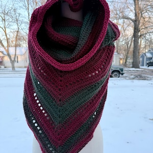 May include: A handmade crochet cowl and hood in shades of burgundy and forest green. The cowl is triangular, with a hood that frames the face. The yarn has a gradient effect, transitioning between colors. The cowl is displayed on a mannequin head.