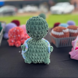 Peut inclure: Un jouet en forme de tortue crocheté de couleur verte avec des accents bleus et blancs. La tortue est posée sur une surface noire.