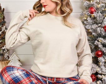 May include: A cream colored sweatshirt with long sleeves and a crew neck. The sweatshirt is worn by a person sitting down, wearing red and blue plaid pajama pants.