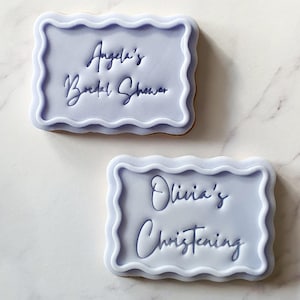 May include: Two decorated sugar cookies with purple icing. The top cookie has the text "Angela's Bridal Shower" and the bottom cookie has the text "Olivia's Christening".