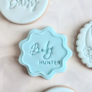 May include: A light blue sugar cookie with a scalloped edge, decorated with a blue icing design that says "Baby Hunter".