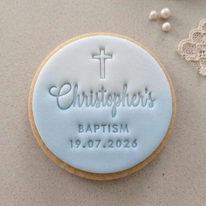 May include: A round biscuit with pale blue icing, embossed with the name "Christopher's", the word "BAPTISM", and the date "19.07.2026". A cross is embossed at the top. The biscuit has a golden-brown edge.