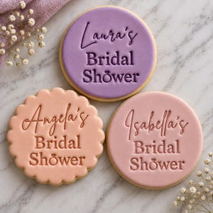 May include: Three round sugar cookies with smooth fondant icing in lavender, peach, and dusty rose. Each cookie features embossed text reading Laura's Bridal Shower, Angela's Bridal Shower, and Isabella's Bridal Shower with a ring icon.
