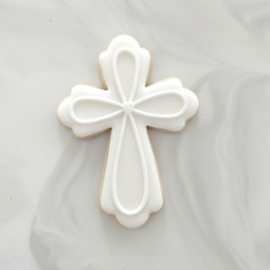 May include: A white iced biscuit in the shape of a cross. The biscuit has a light tan base with white icing details. The icing design features a looped pattern and a central point where the lines meet.