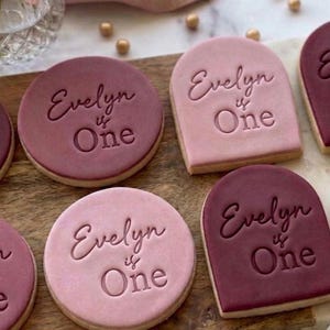 May include: Round and arch-shaped sugar biscuits in shades of dusty rose and deep plum. Each biscuit features the embossed text Evelyn is One in a cursive and sans-serif font combination. The biscuits are arranged on a wooden surface.