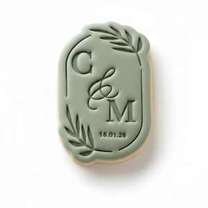 May include: A sage green cookie cutter in an oval shape, featuring the letters "C & M" with an ampersand, and the date "16.01.26". The design includes leaf accents around the edges, ideal for creating personalized biscuits for special events.