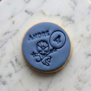 May include: A round biscuit topped with a smooth, matte blue fondant disc. The fondant is embossed with the name ANDRE, a cartoon superhero character, and a circular balloon featuring the number 4, creating a textured, debossed design.