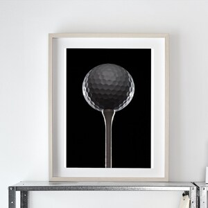 Op de afbeelding: Een witte printer staat op een metalen plank met daarboven een ingelijst zwart-wit foto van een golfbal op een tee. Drie witte kandelaars staan op de plank boven de printer. Een zwarte gieter en een witte klok staan op de plank boven de kandelaars.