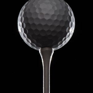Op de afbeelding: Een witte golfbal op een zilveren tee tegen een zwarte achtergrond.