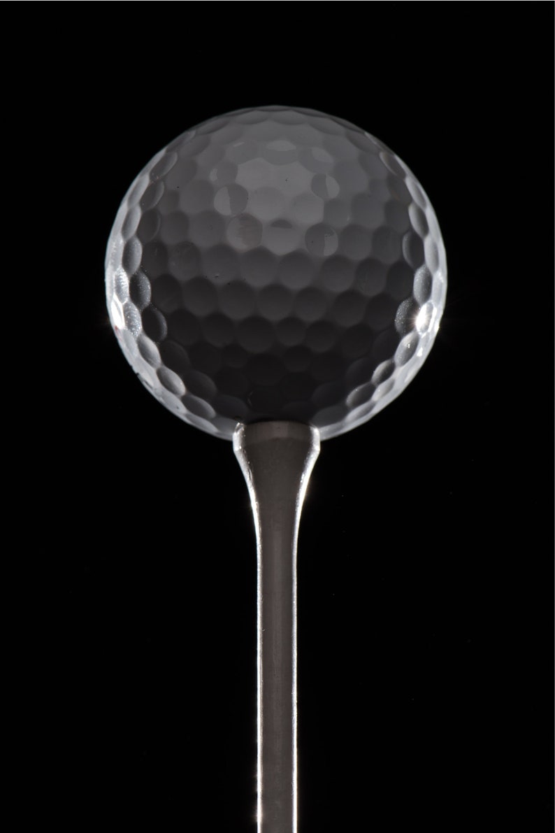 Op de afbeelding: Een witte golfbal op een zilveren tee tegen een zwarte achtergrond.
