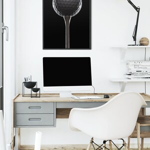 Op de afbeelding: Een zwart-witfoto van een golfbal op een tee, ingelijst en hangend aan een witte muur boven een bureau met een computer, een lamp en een stoel.