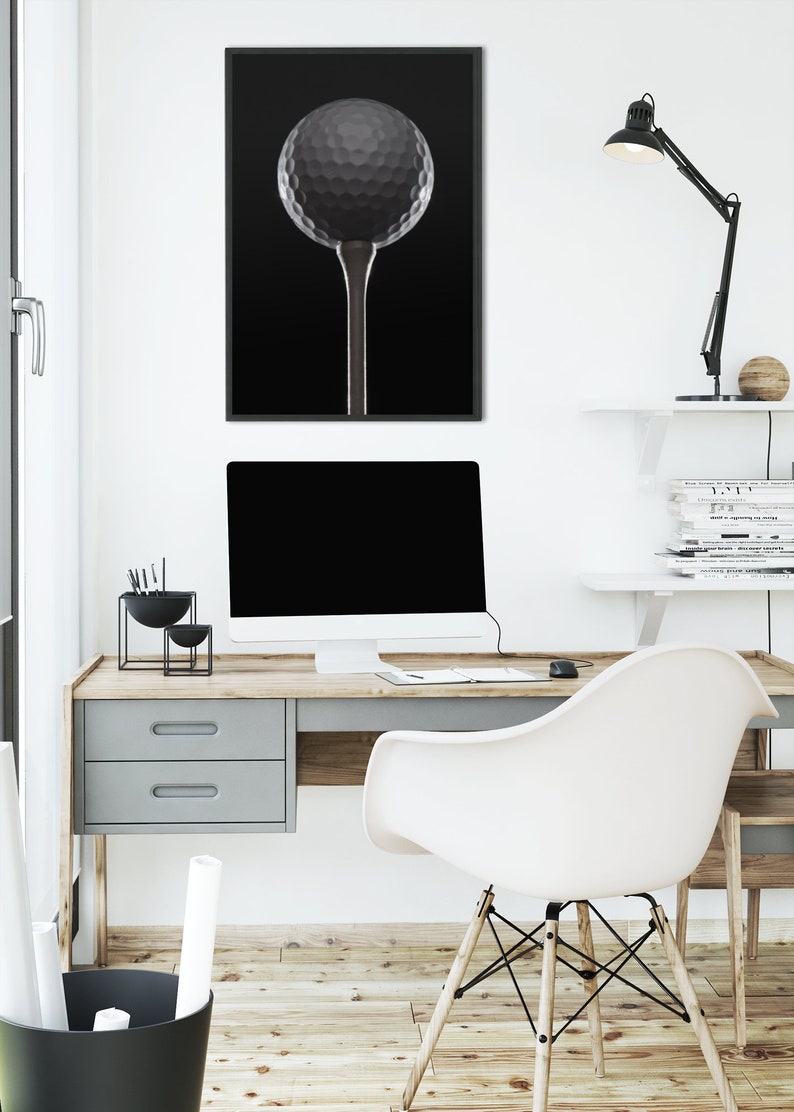 Op de afbeelding: Een zwart-witfoto van een golfbal op een tee, ingelijst en hangend aan een witte muur boven een bureau met een computer, een lamp en een stoel.