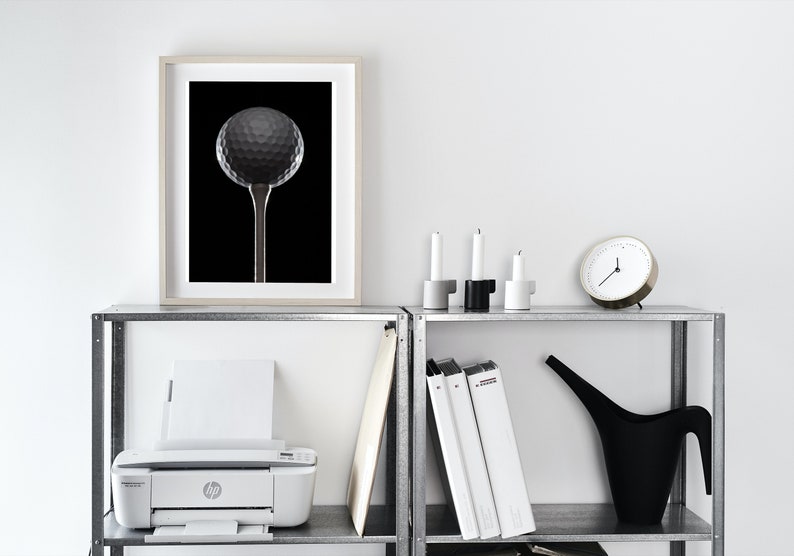 Op de afbeelding: Een witte printer staat op een metalen plank met daarboven een ingelijst zwart-wit foto van een golfbal op een tee. Drie witte kandelaars staan op de plank boven de printer. Een zwarte gieter en een witte klok staan op de plank boven de kandelaars.