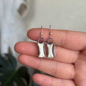 K&ouml;nnte beinhalten: Ein Paar silberne Ohrringe mit wei&szlig;em, rechteckigem Design. Die Ohrringe haben einen strukturierten Rand und einen kleinen Haken f&uuml;r das Ohr. Die Ohrringe werden in einer Hand gehalten.
