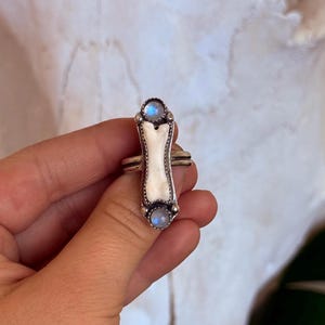 May include: A silver ring with a bone-shaped center and two round, blue-hued stones. The ring has a detailed design with a white bone-like center and is held by a hand against a soft background.
