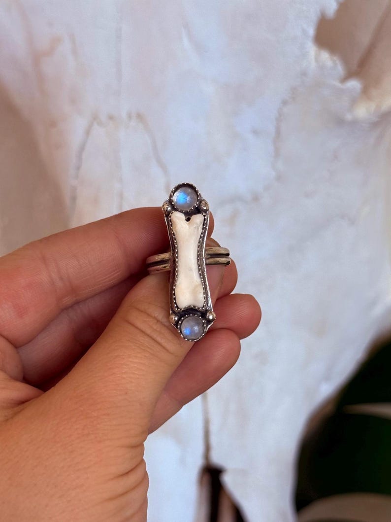 May include: A silver ring with a bone-shaped center and two round, blue-hued stones. The ring has a detailed design with a white bone-like center and is held by a hand against a soft background.
