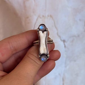 May include: A silver ring featuring a bone-shaped centerpiece with a white inlay. The ring is adorned with two round, light blue moonstone cabochons, one at each end of the bone design. The ring has a double band.