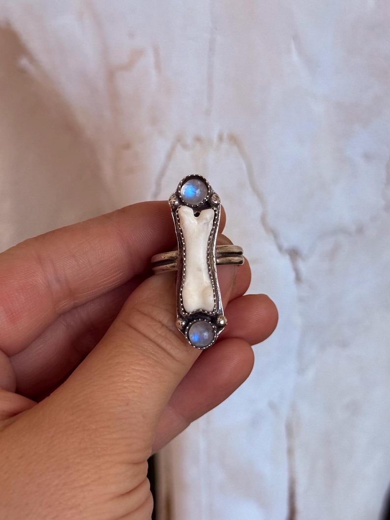 May include: A silver ring featuring a bone-shaped centerpiece with a white inlay. The ring is adorned with two round, light blue moonstone cabochons, one at each end of the bone design. The ring has a double band.