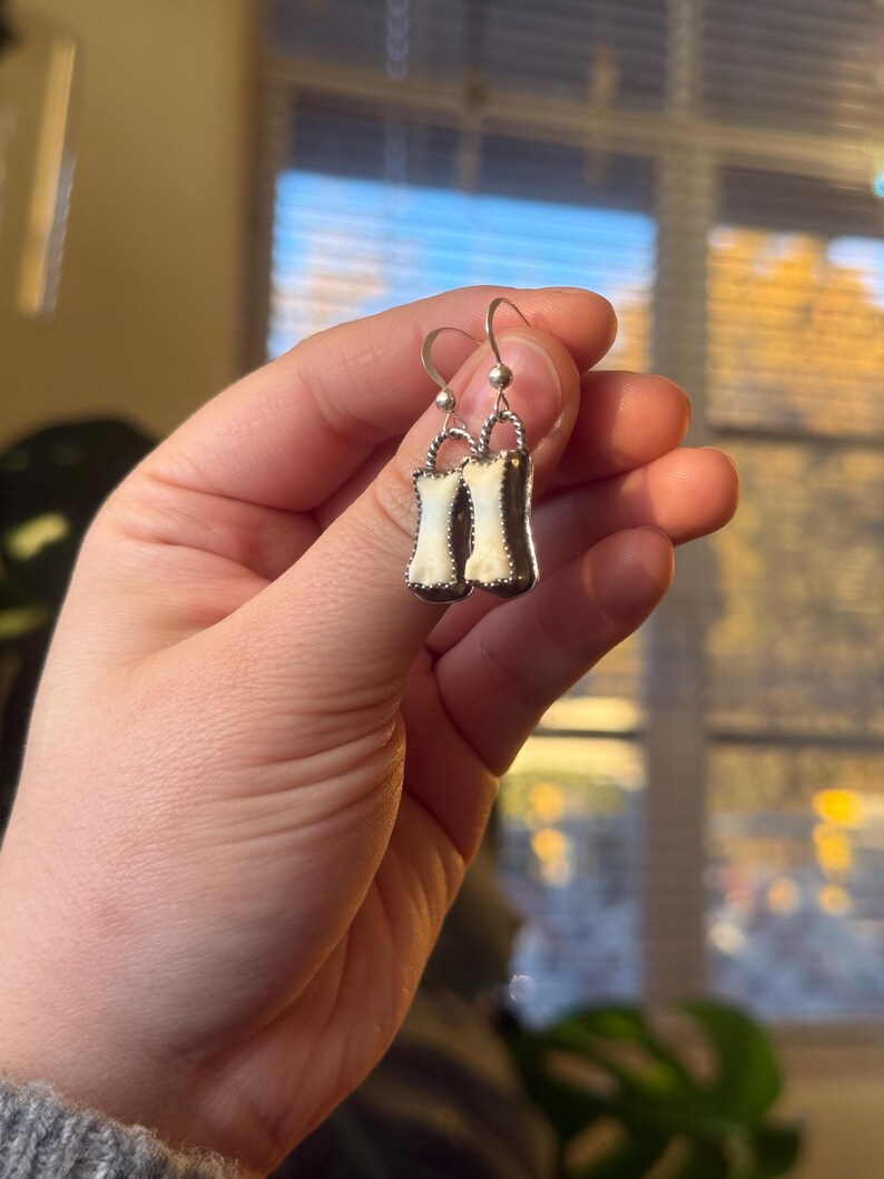 K&ouml;nnte beinhalten: Silberfarbene Ohrringe mit Korsett-Design. Jeder Ohrring hat eine wei&szlig;e Mitte und einen dunklen Rand, mit einem kleinen silbernen Ball und Haken f&uuml;r das Ohr. Die Ohrringe werden von einer Hand gehalten.