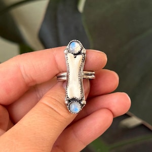 May include: A silver ring featuring a bone-shaped centerpiece with a white, textured surface. Two small, round, blue-toned gemstones are set at the top and bottom of the bone. The ring has a double band design.